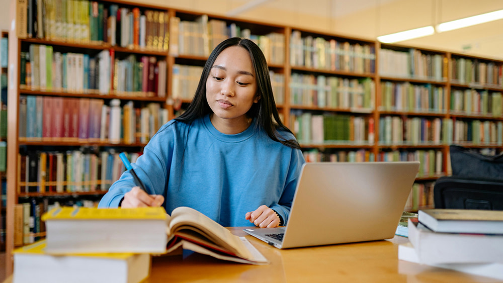 A female international student sits at a table with a stack of books in her US university library, doing research for an upcoming term paper.