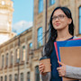 A female international student holds notebooks and a cup of coffee in front of a university hall on campus in the USA