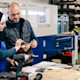 A female international student wearing a hijab stands in front of a table with an electrical drill, tools, and papers and works on a hands-on mechanical engineering project with her professor at a US university