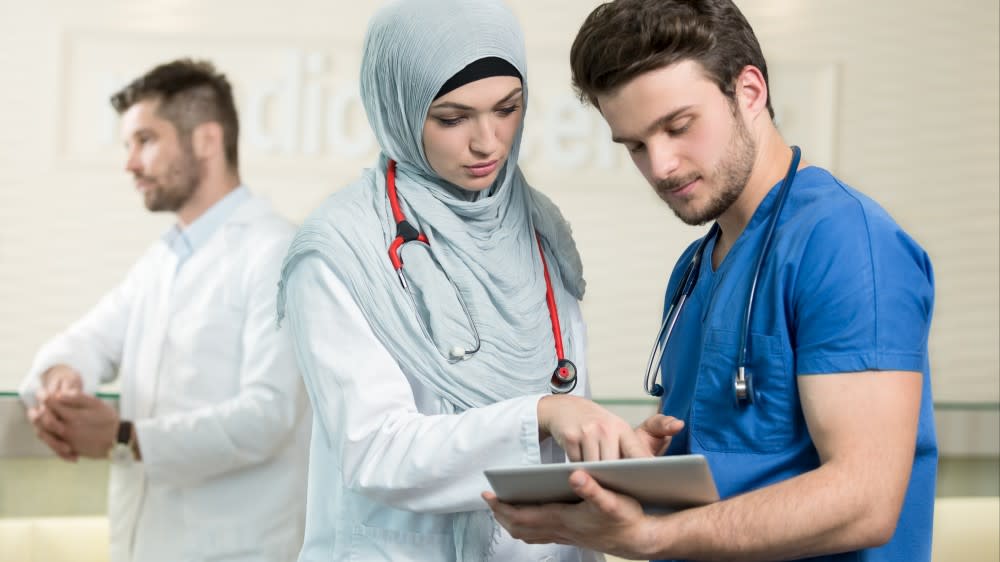 A young female doctor wearing a hijab confers with a young male doctor in scrubs.