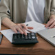 This photo shows the hands of a future accountant working on accounting spreadsheets with a calculator, laptop, and notebook as she does homework for her accounting courses at a US university.