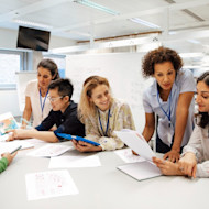 Five employees wearing company lanyards seated at a table, collaborating on work with a whiteboard in the background.