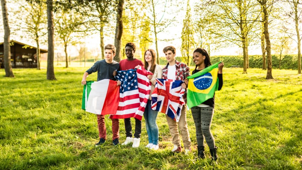 A diverse group of five international students stand in a field and hold flags from Italy, the USA, the UK, and Brazil.