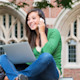 A female international student sits outdoors on a US college campus with a laptop on her lap while talking on a cell phone
