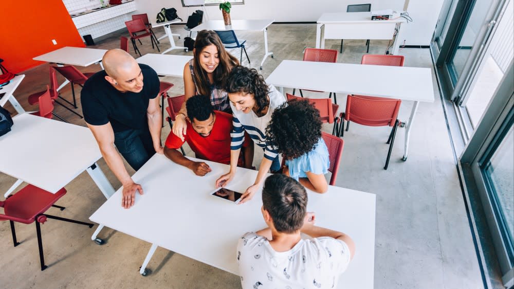 Five students gather around a tablet computer at a desk in a classroom.