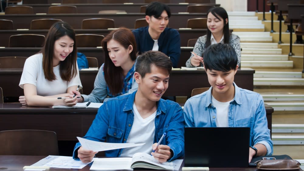 Two students sitting a lecture hall while looking at a laptop, with three other students seated behind them.
