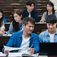 Two students sitting a lecture hall while looking at a laptop, with three other students seated behind them.