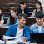 Two students sitting a lecture hall while looking at a laptop, with three other students seated behind them.