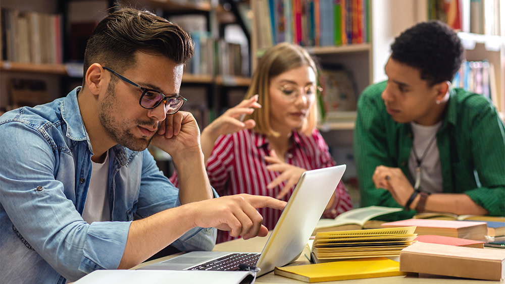 A group of three Canadian students sit at a table with laptops preparing their student visa USA from Canada.