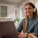 A female international student wearing headphones sits at a table in front of a laptop for her online class.