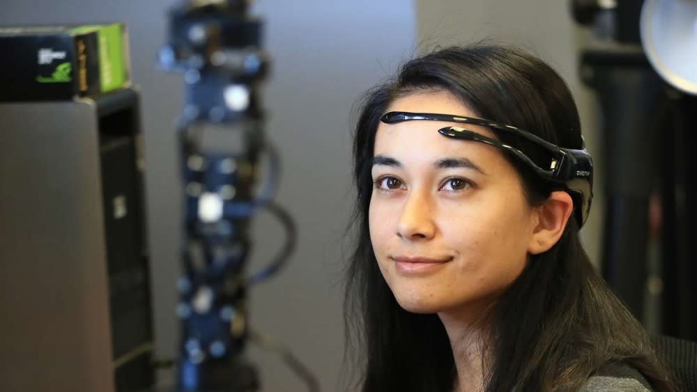 A UDayton female international student engineer sits in front of an AI tower device wearing head gear