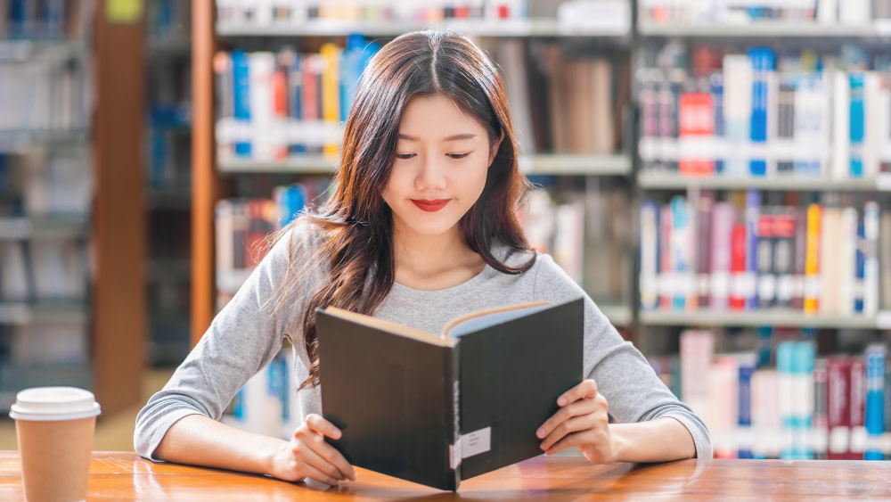 A female Asian international student sits in her US university library and reads an English book with a cup of coffee.