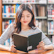 A female Asian international student sits in her US university library and reads an English book with a cup of coffee.