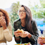 Two women and one man go out for American Food, eating burgers, sitting at a table, with one covering her mouth as she laughs. In American culture food brings many students together in social situations.