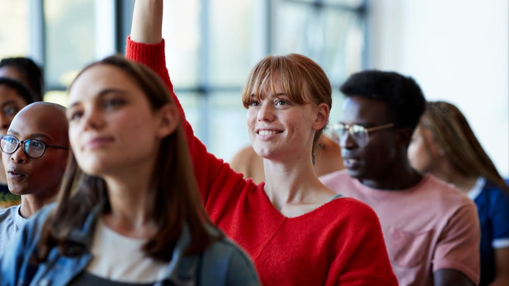 A female international student in a red sweater sits in class and raises her hand to answer a question