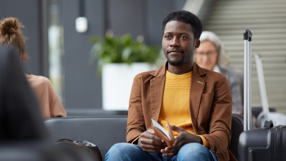 A young Black man sits at an airport holding his passport and student visa with his suitcase next to him