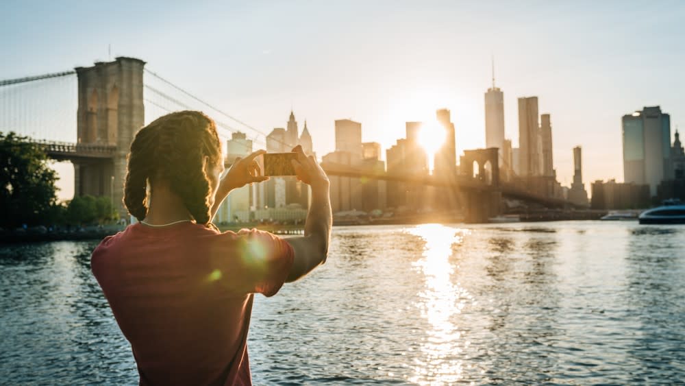An international student stands in front of a river against the Brooklyn Bridge and NYC skyline at sunset taking a photograph.