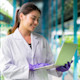 A female international student wearing a lab coat, working toward a food technology specialization for her US university degree, stands with a laptop in a greenhouse near rows of leafy green plants.