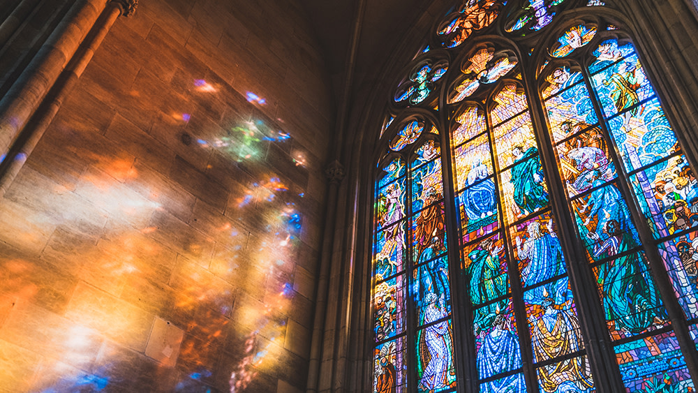 This image shows an oversized stained glass window at a Christian college in the US reflecting colorful sunbeams on an adjacent stone wall.