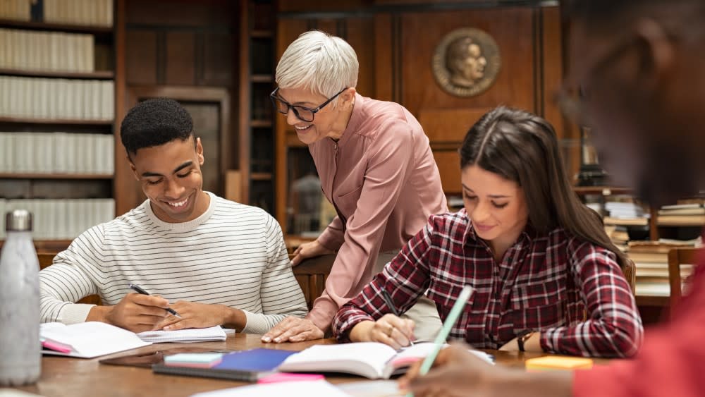 At a table in a university library, a male international student asks his female professor, "what is a thesis?" and she explains the thesis meaning to him looking over his shoulder, with both next to another female student working on her thesis