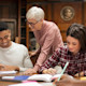 At a table in a university library, a male international student asks his female professor, "what is a thesis?" and she explains the thesis meaning to him looking over his shoulder, with both next to another female student working on her thesis