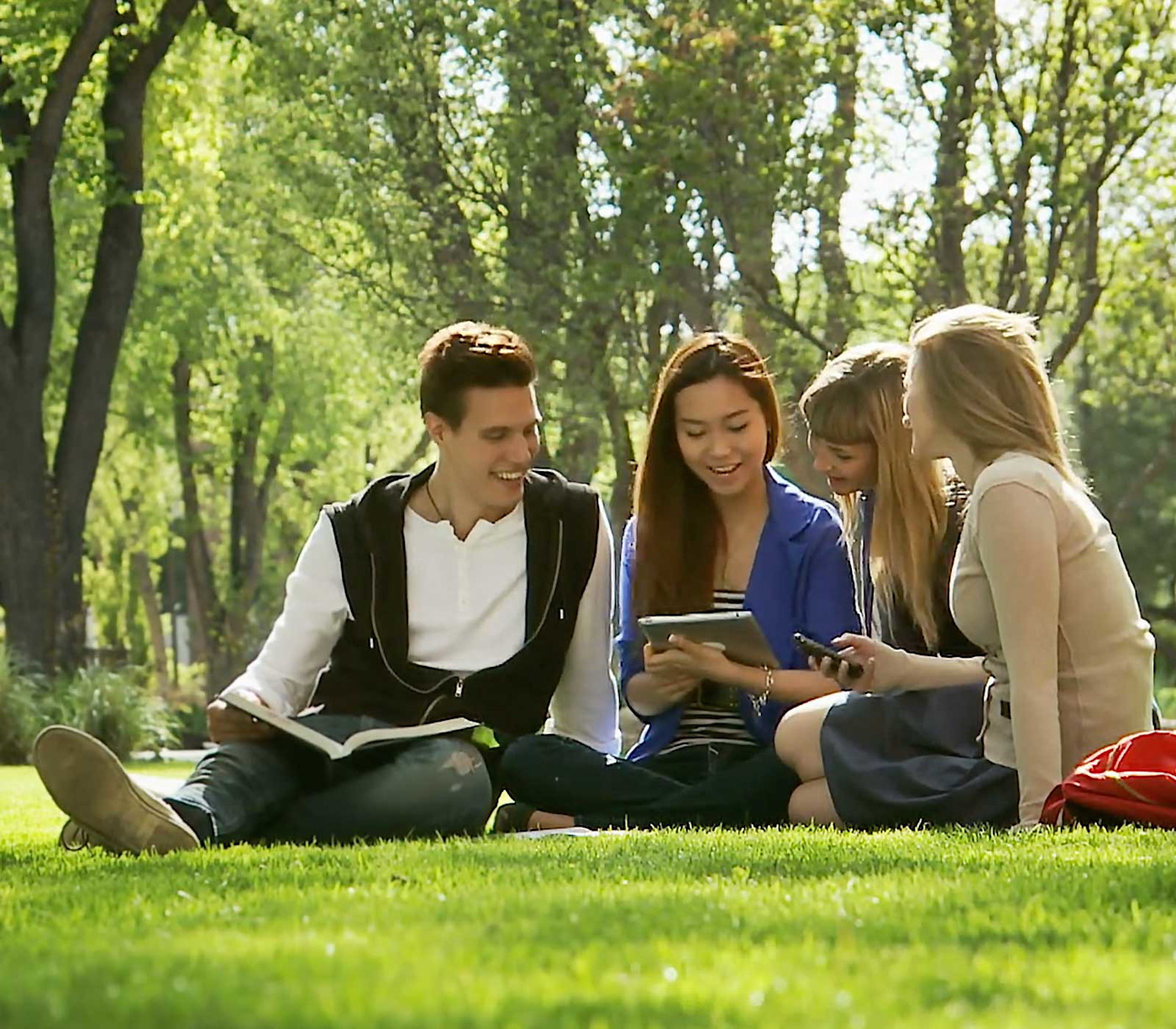 A group of international students sit outside on their college campus quad and look at a book and a tablet