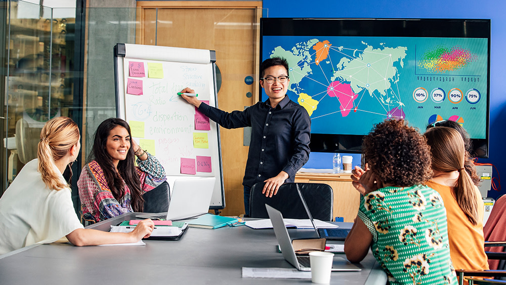 A group of international students sit around a conference table while one student makes a communication skills presentation at a easel in front of a world map