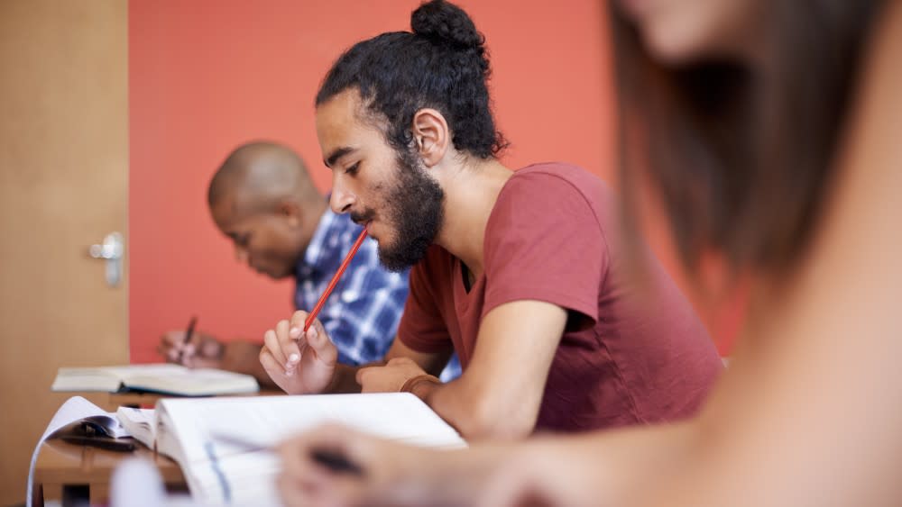 A student with a man bun holds a pencil in his mouth while contemplating an answer to his GRE exam. What is the GRE test? It stands for Graduate Records Examination and is an admissions exam for graduate school degree programs in the US.