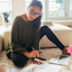 A female international student from Asia wears glasses and sits on the floor in front of a laptop and notebook as she works on a paper before the deadline
