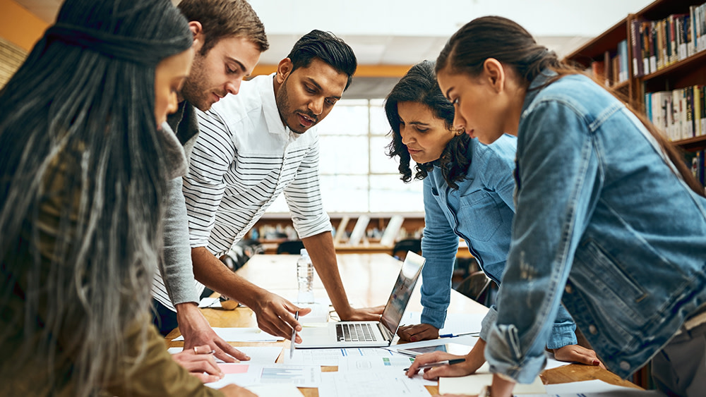 A group of international students in a pathway program at a US university stand around a classroom table with an open laptop and papers as their professor goes over an assignment.
