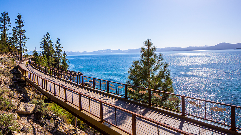 In the Reno area, a pedestrian and biking walkway borders the shores of Lake Tahoe with the Sierra mountains in the background and evergreen trees in the foreground