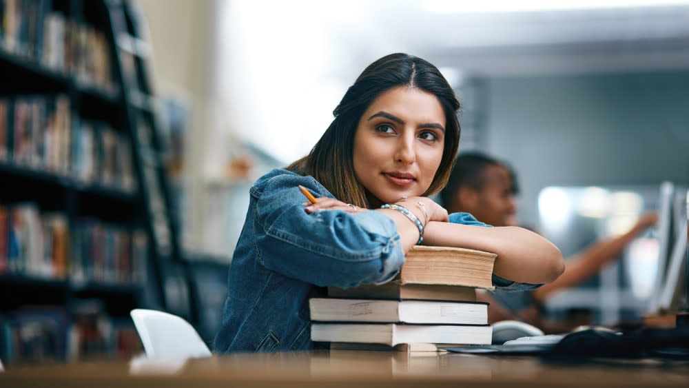 In considering how to get a student visa USA from India, a female Indian student sits with her arms folded on a stack of books in a library to research student visa requirements for USA and determine how to get a u.s. student visa.