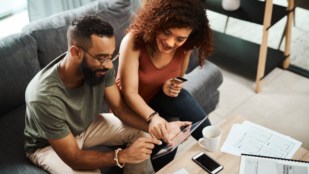 A man and woman discussing what's on a tablet while sitting on a sofa in front of a coffee table.