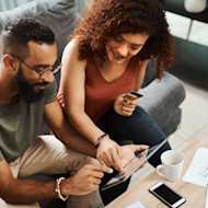 A man and woman discussing what's on a tablet while sitting on a sofa in front of a coffee table.