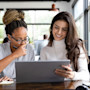 Two female international students sit side by side in front of a laptop