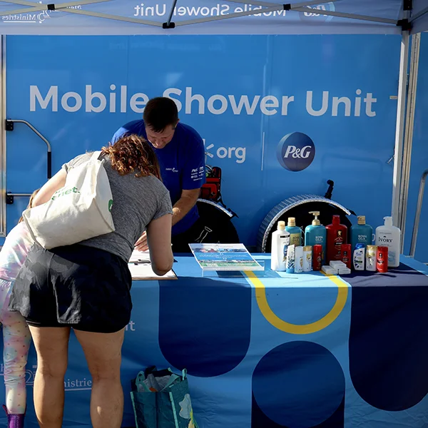A woman and her young daughter stand in front of a bright blue table with a volunteer, filling out a form to use the mobile shower unit. P&G personal care products are displayed on the table.