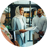 A smiling woman wearing a light-colored hijab holds a stack of papers, surrounded by an engaged group of people in an office environment. A blue circle outline surrounds the icon.