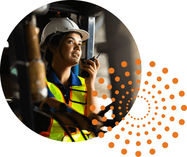 A worker with a white hard hat and high-visibility safety vest talks into a walkie-talkie is in an industrial setting. In the lower right hand corner, there is a pattern of orange dots creating five concentric circles.