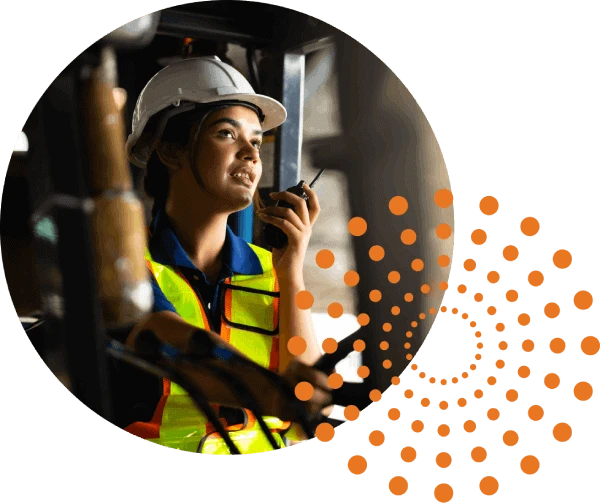 A worker with a white hard hat and high-visibility safety vest talks into a walkie-talkie is in an industrial setting. In the lower right hand corner, there is a pattern of orange dots creating five concentric circles.