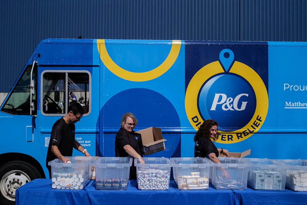 Volunteers sort supplies in front of a blue P&G Disaster Relief truck at a community support event.