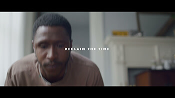 A Black man with short, dark hair. He wears a brown t-shirt. White text in the center says, "reclaim the time."