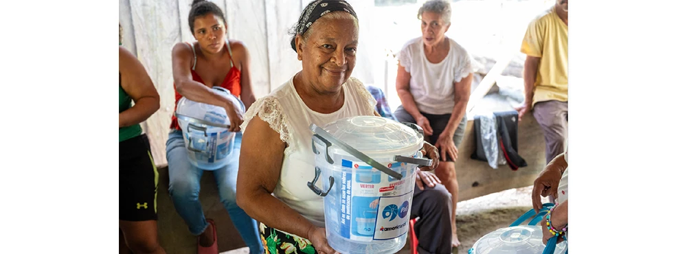 A woman smiling at the camera holds a bucket from the CSDW program, which helps turn contaminated water into clean drinking water. Two other women, one with a bucket, are visible in the background.