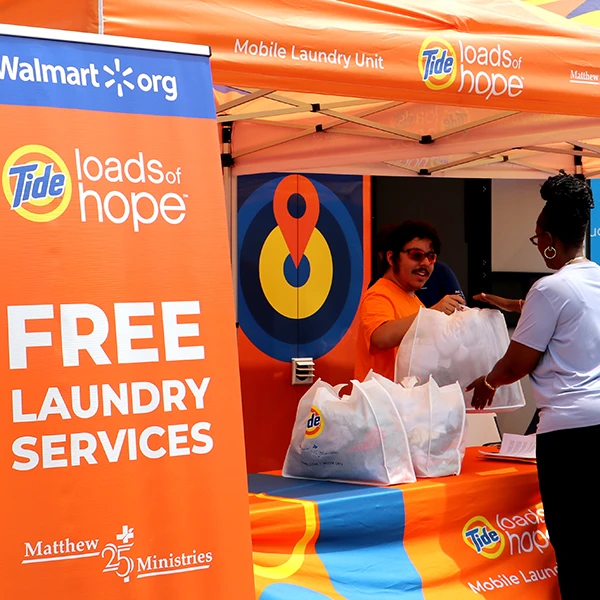 A man behind a Tide Loads of Hope orange table and tent hands a woman a Tide-branded bag of clean clothes, with a large sign that says "Free Laundry Services."