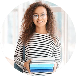A young woman with long, brown curly hair and glasses smiles directly at the camera. She is wearing a black and white striped shirt and is holding a set of books.