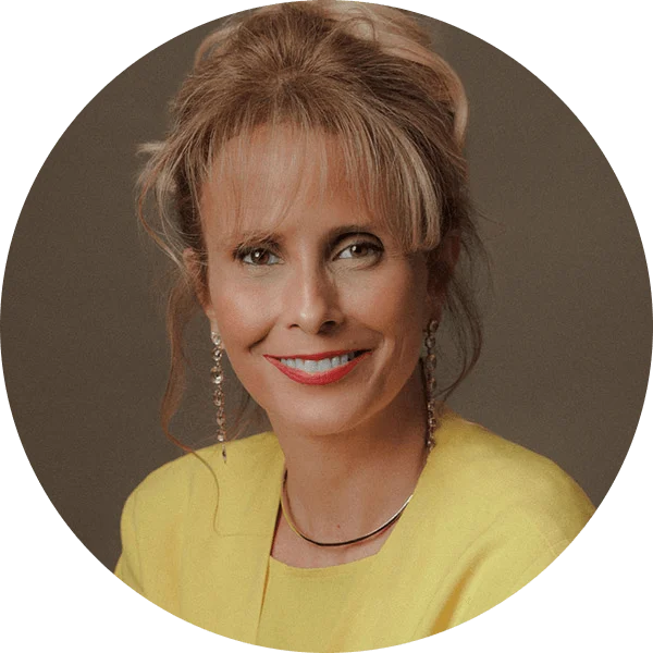 A woman with blonde hair smiles into the camera. She is wearing a yellow shirt and sweater set.