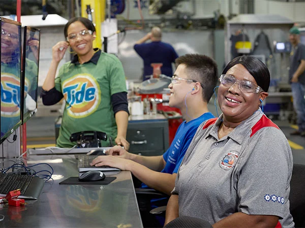 Two women and a man sit in front of several computers in a manufacturing plant. They wear protective eye gear and smile at the camera.
