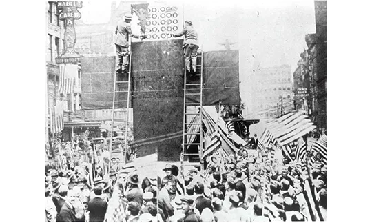 A 1917 black and white photo of two men on ladders painting a sign for a Red Cross donation drive in Cincinnati’s Fountain Square, with a crowd of onlookers, some holding American flags.