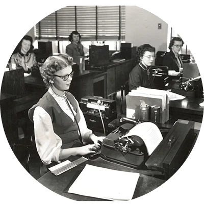 A black and white photo of P&G’s first blind employee, Jane Dotson, who was hired in January 1950 and worked in the transcribing department at the General Offices. She sits at a desk, typing on a typewriter amongst other women transcribers.