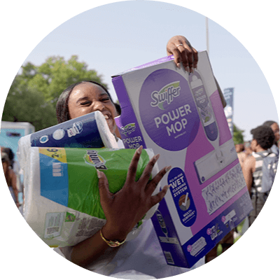 A young Black woman smiles as she carries an armful of household products, such as paper towels and toilet paper.