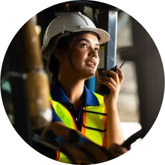A worker with a white hard hat and high-visibility safety vest talks into a walkie-talkie is in an industrial setting.